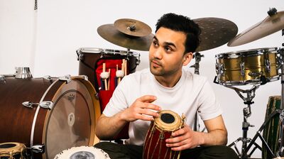 A man in a white t-shirt sits on the floor surrounded by a drum kit and several traditional hand drums.