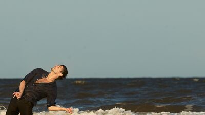 A wet man in a dark button-down shirt arches his back, dancing dramatically in the ocean surf against a clear sky.