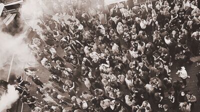 High-angle sepia-toned photo of a large, dense crowd gathering outdoors near a stage with smoke effects.