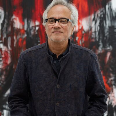 Artist Anish Kapoor stands in front of a red, white and black background. His hands are in the pocket of his brown coat and he looks directly at the camera
