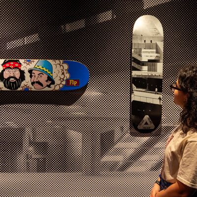 A woman looks at skateboard decks mounted on a polka-dot wall, featuring Cheech & Chong graphics and architectural photography.