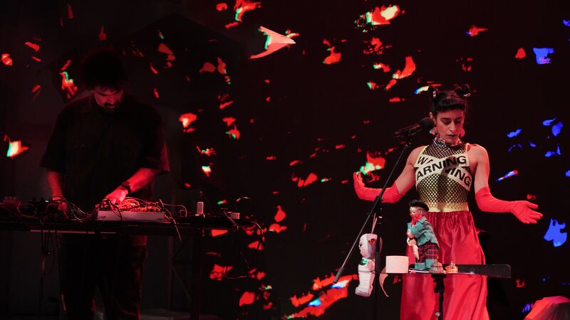 Two musicians perform on a dark stage with red light projections; the singer wears red gloves and "Warning" tape.