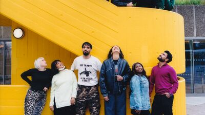 A diverse group of twelve people pose playfully on and underneath a bright yellow external concrete staircase outdoors.