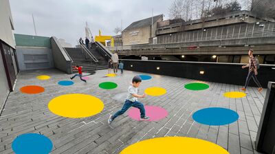 A young boy jumps across large colorful dots painted on the ground at London's Southbank Centre on a sunny day.