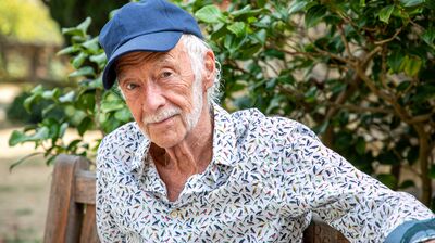 Roger McGough, an elderly White man, seated on a wooden bench wearing a blue cap