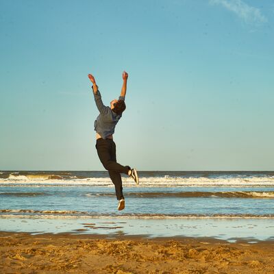 A person in casual clothes leaps high into the air with arms reaching up on a sunny beach with gentle waves.