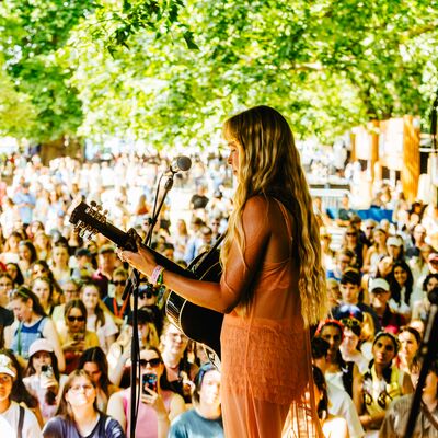 A blonde woman plays an acoustic guitar on stage, facing a massive outdoor festival crowd under a canopy of green trees.