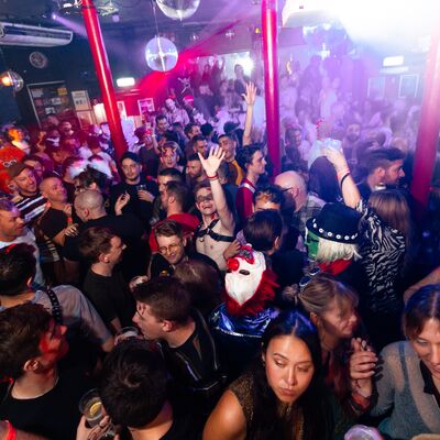 A packed, vibrant nightclub crowd dances under purple lights and disco balls, with some people in festive costumes.