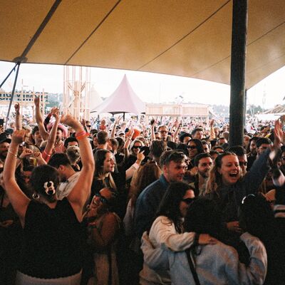 A large, joyful crowd dances with their arms raised under a tan canopy at an outdoor music festival during the day.