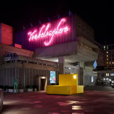 A brutalist building at night, featuring a large pink neon sign that reads "You belong here" above a yellow structure.