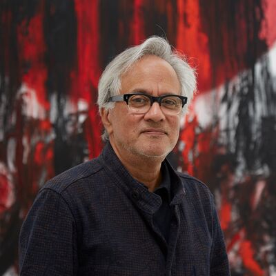 Anish Kapoor wearing glasses and a black suit, standing in front of a red and black artwork