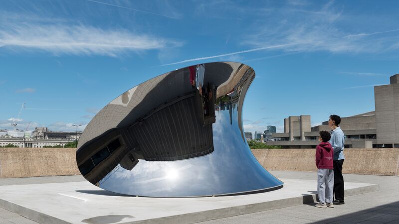 An outdoor wide shot shows two people standing on a concrete terrace looking at a large, curved, mirror-polished steel sculpture that reflects the sky and surrounding brutalist architecture.