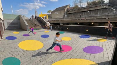 A young boy jumps across large colorful dots painted on the ground at London's Southbank Centre on a sunny day.