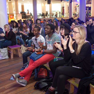 A diverse audience sits on wooden benches in a large indoor hall, smiling and clapping after a performance.