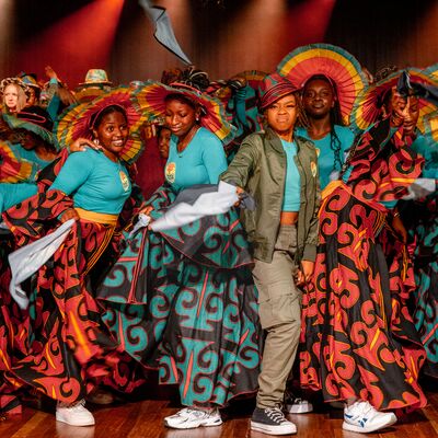 Dancers in teal tops and patterned skirts perform with colorful pleated hats on a stage lit by warm red spotlights.
