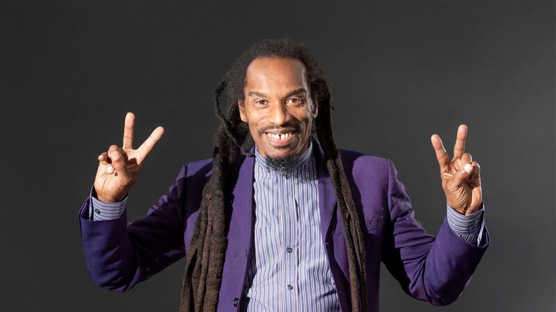 A man with long dreadlocks and a gap-toothed smile wearing a purple suit and making peace signs with both hands.