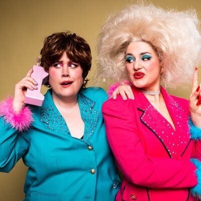 Two performers stand next to each other, one in a blue blazer holding a telephone, and another in a pink blazer