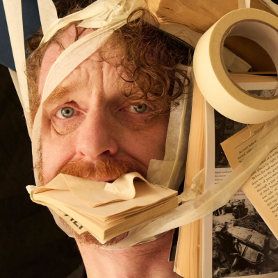 a man in a black blazer looking directly at the camera while a chaotic cluster of books and a roll of masking tape are bound tightly to his head and stuffed into his mouth using beige tape.
