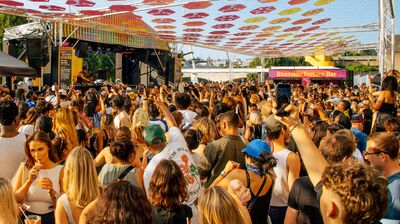 A crowded outdoor music festival with people cheering and holding up phones, beneath a ceiling of orange, yellow and red discs