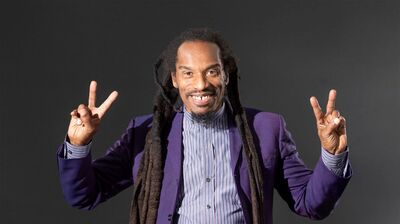 A man with long dreadlocks and a gap-toothed smile wearing a purple suit and making peace signs with both hands.