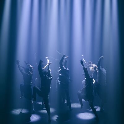 Dancers in silhouette perform on a dark stage beneath a row of intense vertical blue spotlights.