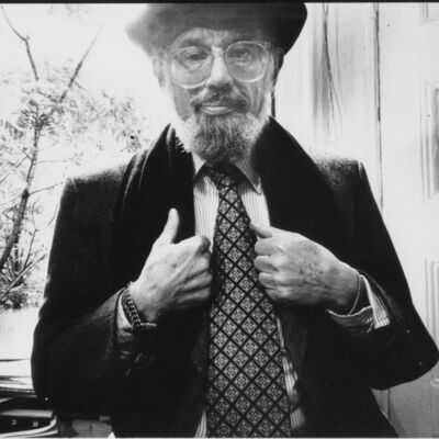 Black and white portrait of a bearded man with glasses and a hat, adjusting his patterned tie while looking at the camera.