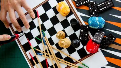A collage of game pieces including chess, dominoes, and pick-up sticks, with a hand using a craft knife to cut a board.