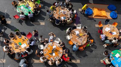 An aerial view of people sitting at several round orange and green tables during an outdoor communal workshop.