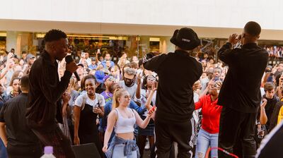 Three performers on an outdoor stage sing into microphones for a large, diverse crowd of people dancing and cheering.