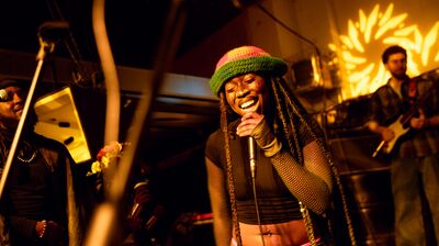 A woman with long dreadlocks and a colorful hat sings into a microphone on stage with a band.