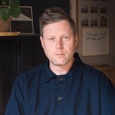 Max porter wears a blue workman's jacket buttoned up to the collar and looks straight at camera. In the background are polaroid images and sketchings.