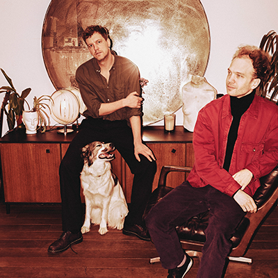Two men pose in a living room with a dog; one sits on a sideboard and the other in a chair, with a large mirror behind.