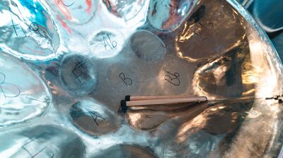 Close-up of a silver steelpan drum with hand-written musical notes and two wooden mallets resting inside.