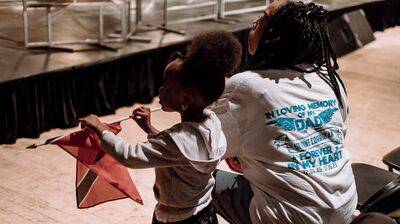 A woman in a memorial t-shirt sits with a young girl holding a Trinidad and Tobago flag, watching an indoor stage performance.ets resting inside.