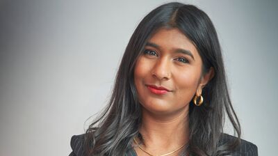 A woman with long dark hair and red lipstick poses in a pinstripe blazer and gold hamsa necklace against a gray wall.