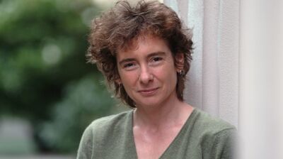 A close-up portrait of a woman with short, curly brown hair wearing a green v-neck sweater, leaning against a white column.