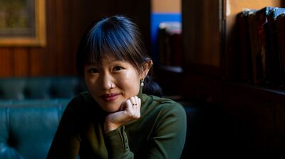Rebecca F Kuang sat at a table in a restaurant, resting her head on her hand with a glass of water next to her.