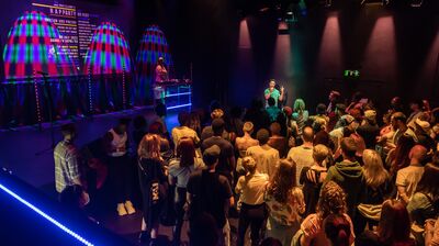 An audience watching a poet with a DJ off to one side, in a studio theatre with multi-coloured lighting
