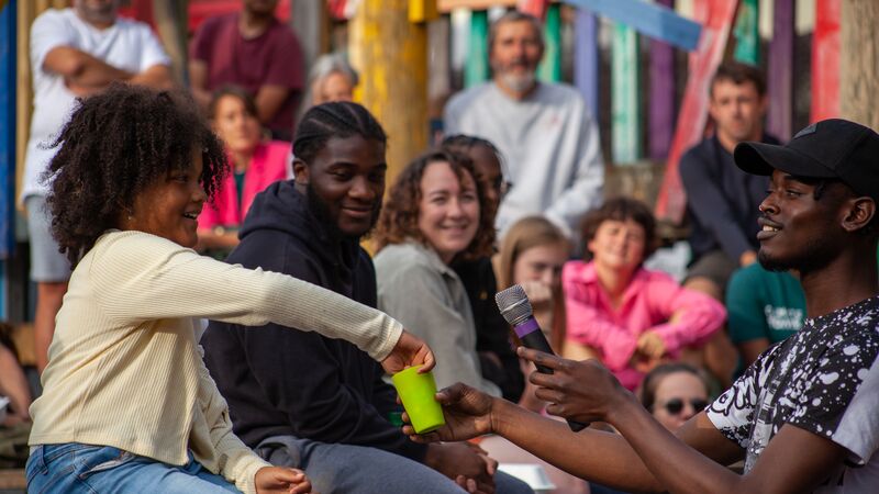 A young girl drops a token into a green cup held by a man with a microphone, while a smiling crowd watches in the background.