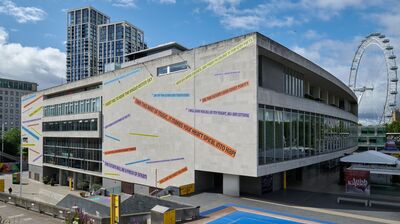 A large concrete building with colourful text decals affixed to its facade stands near a riverfront square, with the London Eye visible in the background.