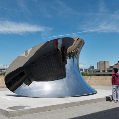 An outdoor wide shot shows two people standing on a concrete terrace looking at a large, curved, mirror-polished steel sculpture that reflects the sky and surrounding brutalist architecture.