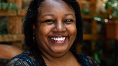 Portrait of author Malorie Blackman. She sits in a garden wearing a patterned blue top and a lace cover up.