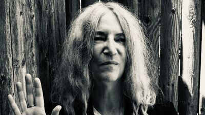 A black and white portrait of Patti Smith raising her hand with palm facing out, leaning against a wooden fence.