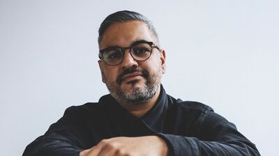 British author and screenwriter Nikesh Shukla wearing a black shirt and leaning on the back of a chair