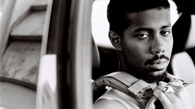 A black and white close-up portrait of a man looking over his shoulder while seated inside a vehicle, gripping a backpack strap.