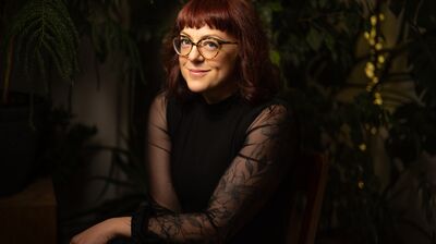 A woman with reddish-brown hair and glasses smiles while sitting in a dimly lit room surrounded by houseplants.