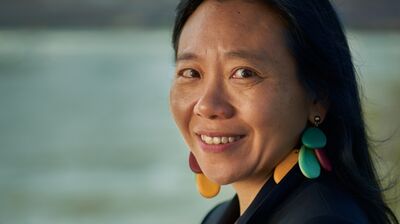A three-quarter portrait of a smiling woman looking back over her shoulder, wearing a dark blazer and colorful earrings.