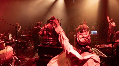 Musicians perform on a stage lit by red spotlights as a crowd cheers in the background.