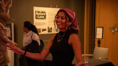 A smiling woman with a colorful headscarf and gold jewelry dances in an indoor space with a "Nature + Love" sign behind.
