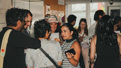 A diverse group of people talks and takes notes in a crowded room with posters on a corkboard in the background.